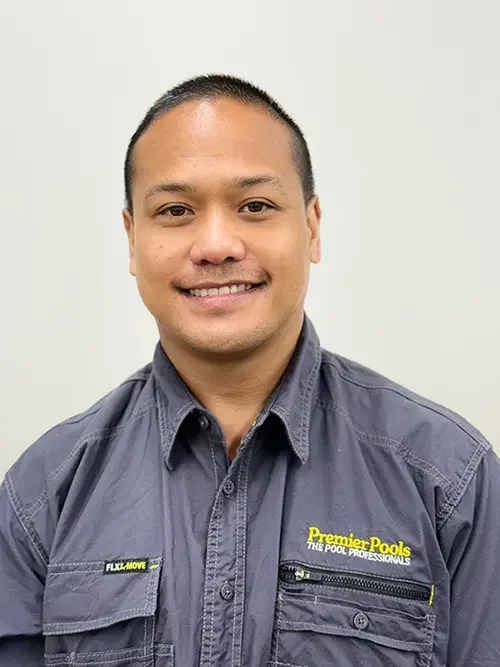 Portrait of a smiling man with short black hair wearing a dark gray work shirt with yellow and white text and zippers, standing against a plain light background.