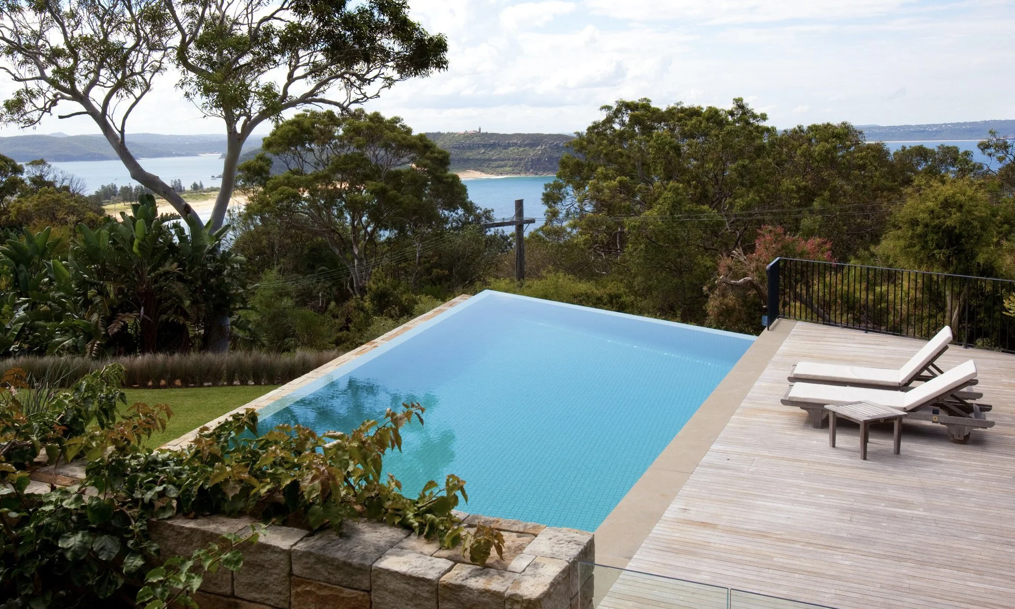 Infinity pool overlooking a lush green landscape with trees and a body of water in the distance, with two lounge chairs on a wooden deck.
