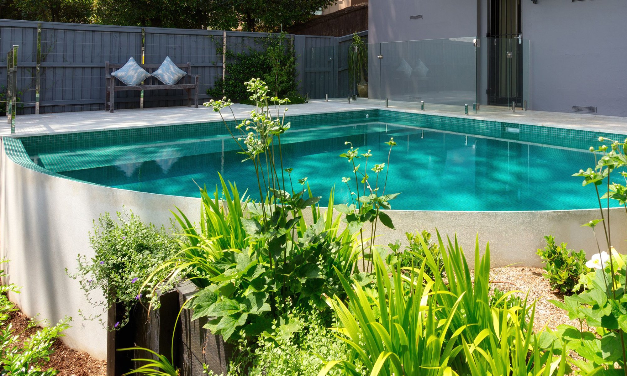 Backyard swimming pool surrounded by greenery and a gray fence, with a seating area and plants in the foreground.