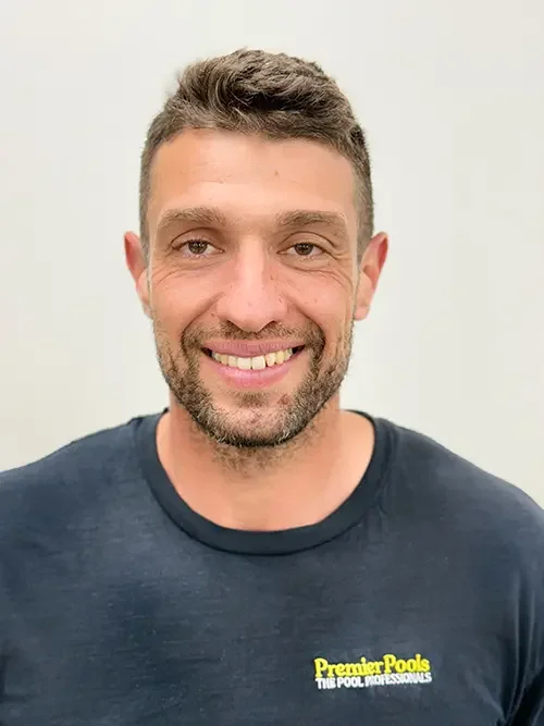 A smiling man with short brown hair, facial hair, and blue eyes, wearing a black T-shirt with yellow and white text that says "Premier Pools The Pool Professionals" on a plain light-colored background.