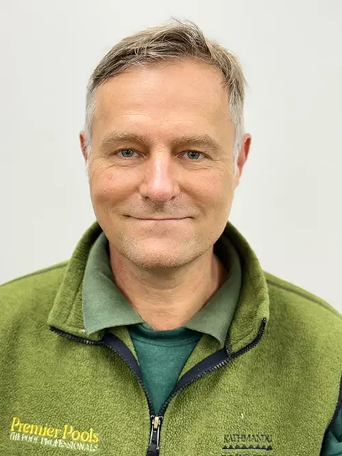 A man with short, light brown hair and blue eyes smiling at the camera, wearing a green pullover with a logo and a green collared shirt underneath.