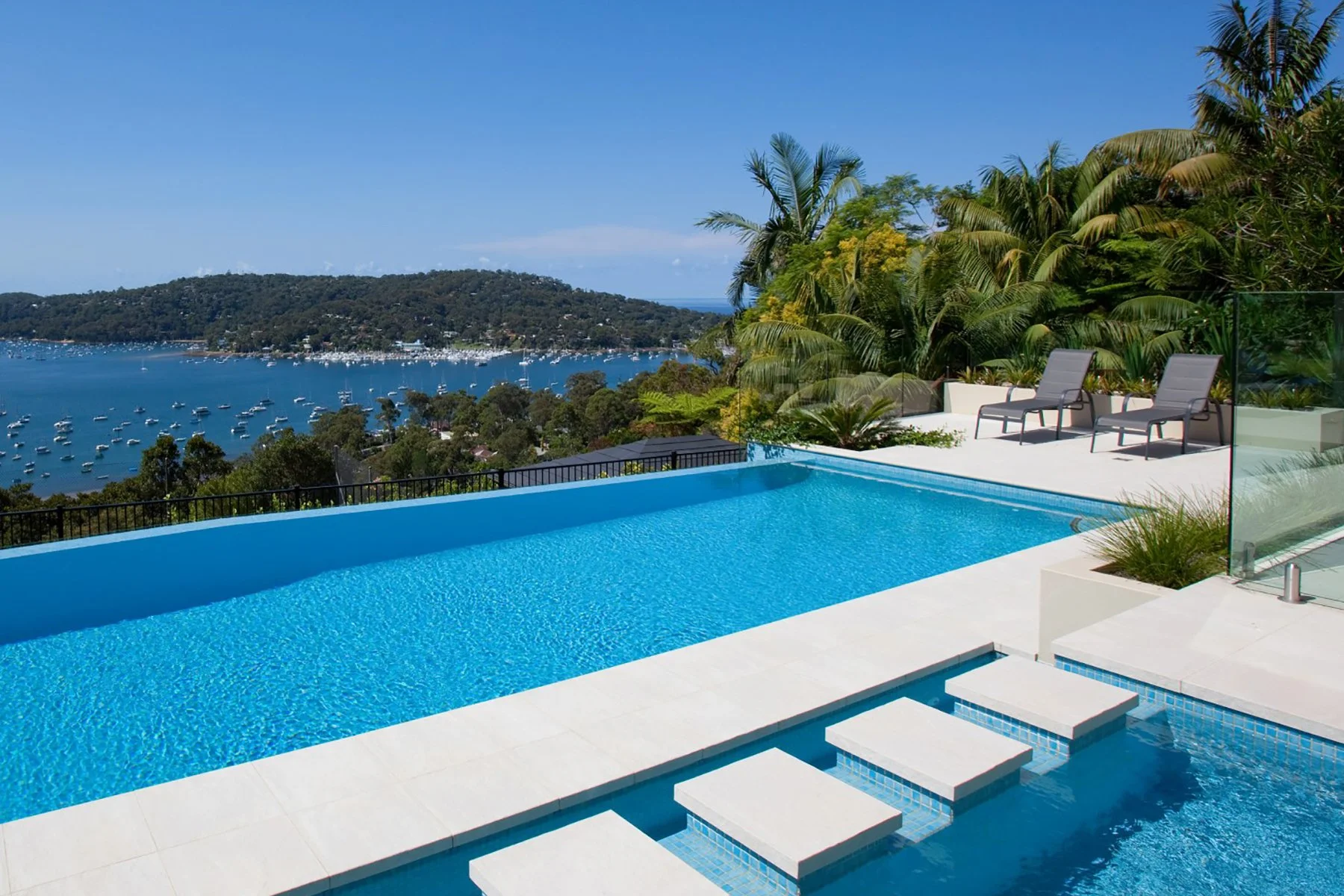 Luxury infinity pool overlooking a bay with boats, surrounded by lush tropical plants and outdoor lounge chairs.