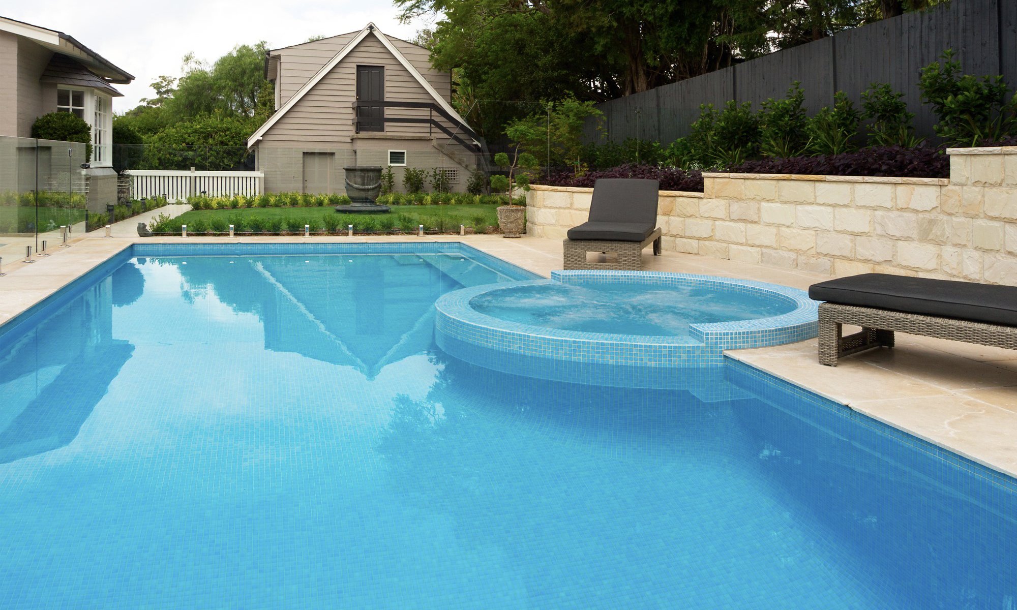 Residential backyard with a swimming pool and hot tub, lounge chairs, lush landscaping, and a house in the background.