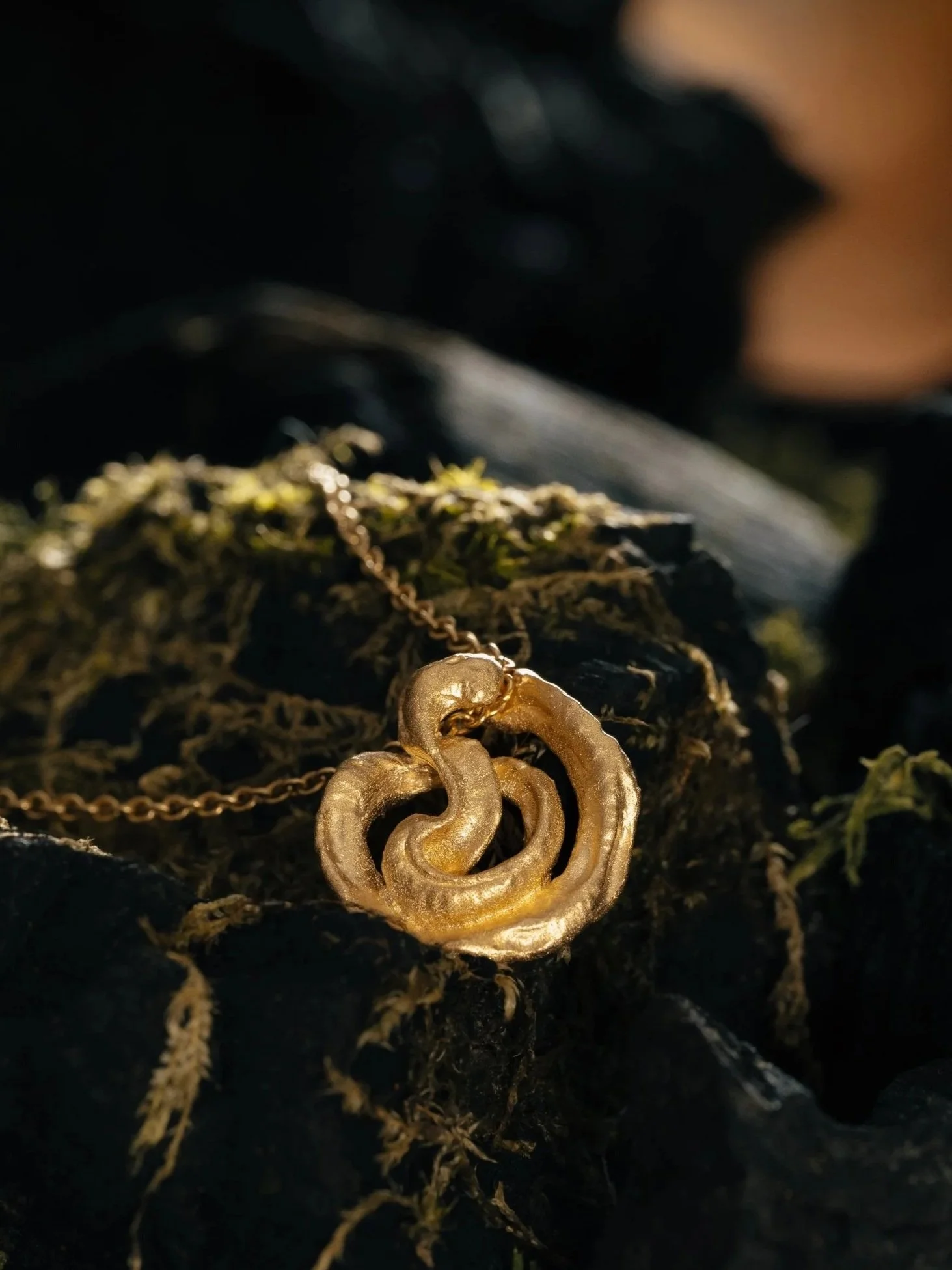 Close-up of a gold heart-shaped locket necklace with intricate design, resting on dark mossy ground.