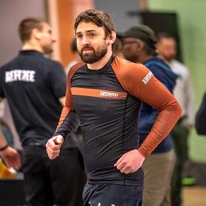 A man in athletic clothing, with a beard, standing indoors with others in the background.