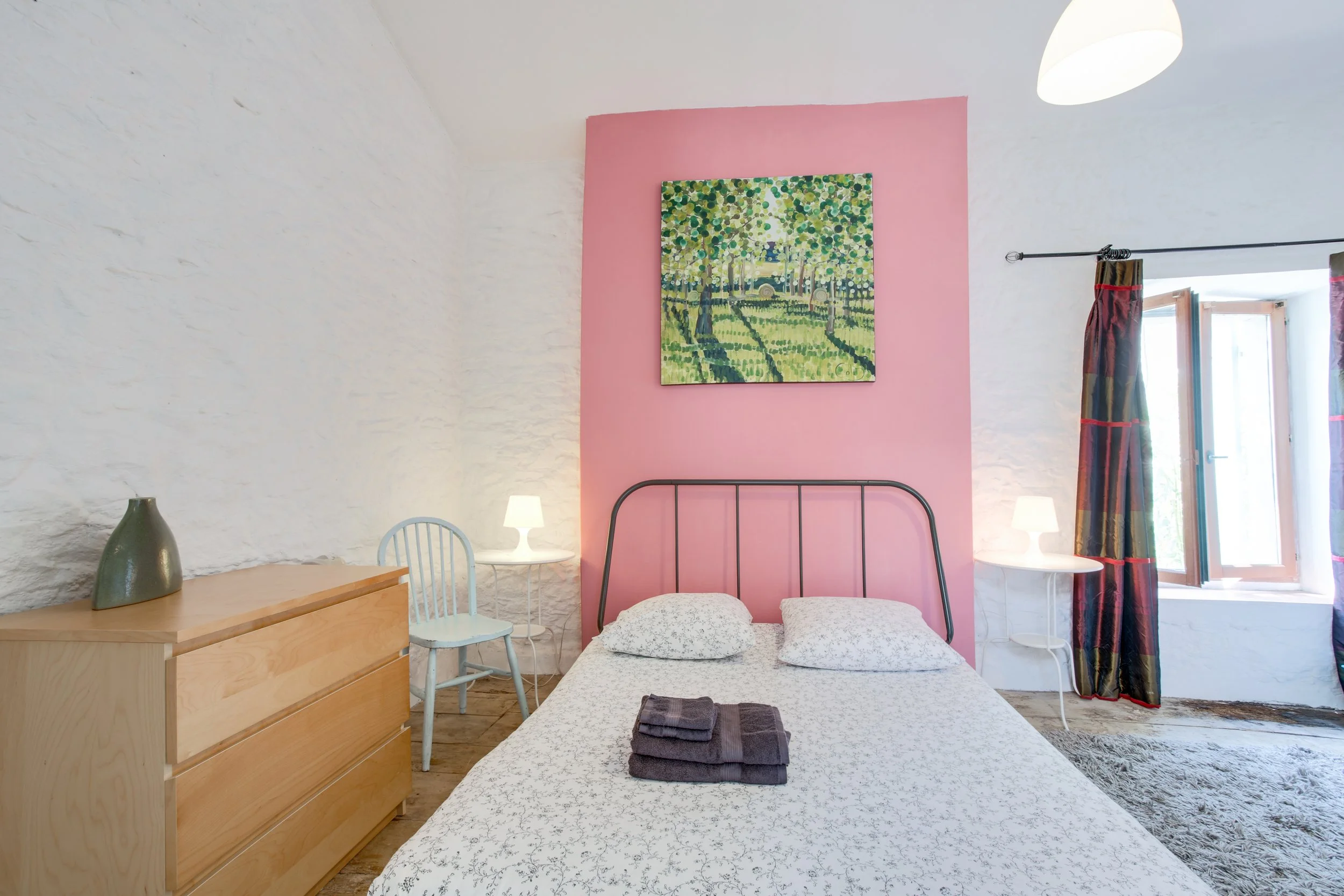 Cozy bedroom with white textured walls, a bed with white patterned bedding, twin pillows, and folded dark towels on it. A pink accent wall behind the bed features a landscape painting. There are small white bedside tables with lamps, a light blue chair, a wooden dresser, and a window with colorful curtains.