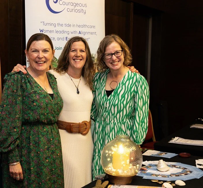 Three women smiling and standing together at an indoor event, with a banner behind them that reads 'Courageous Curiosity' and includes text about healthcare and women. There is a table with decorative items and papers in front of them.