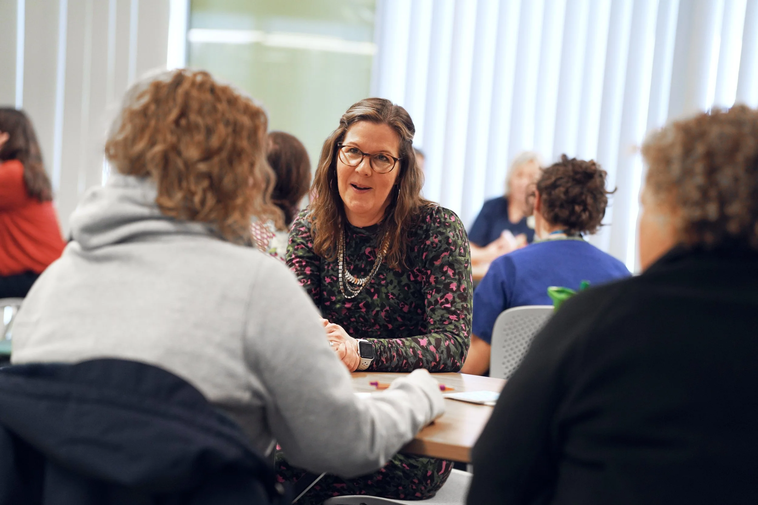A woman with glasses and long brown hair speaking to a group in a meeting room.