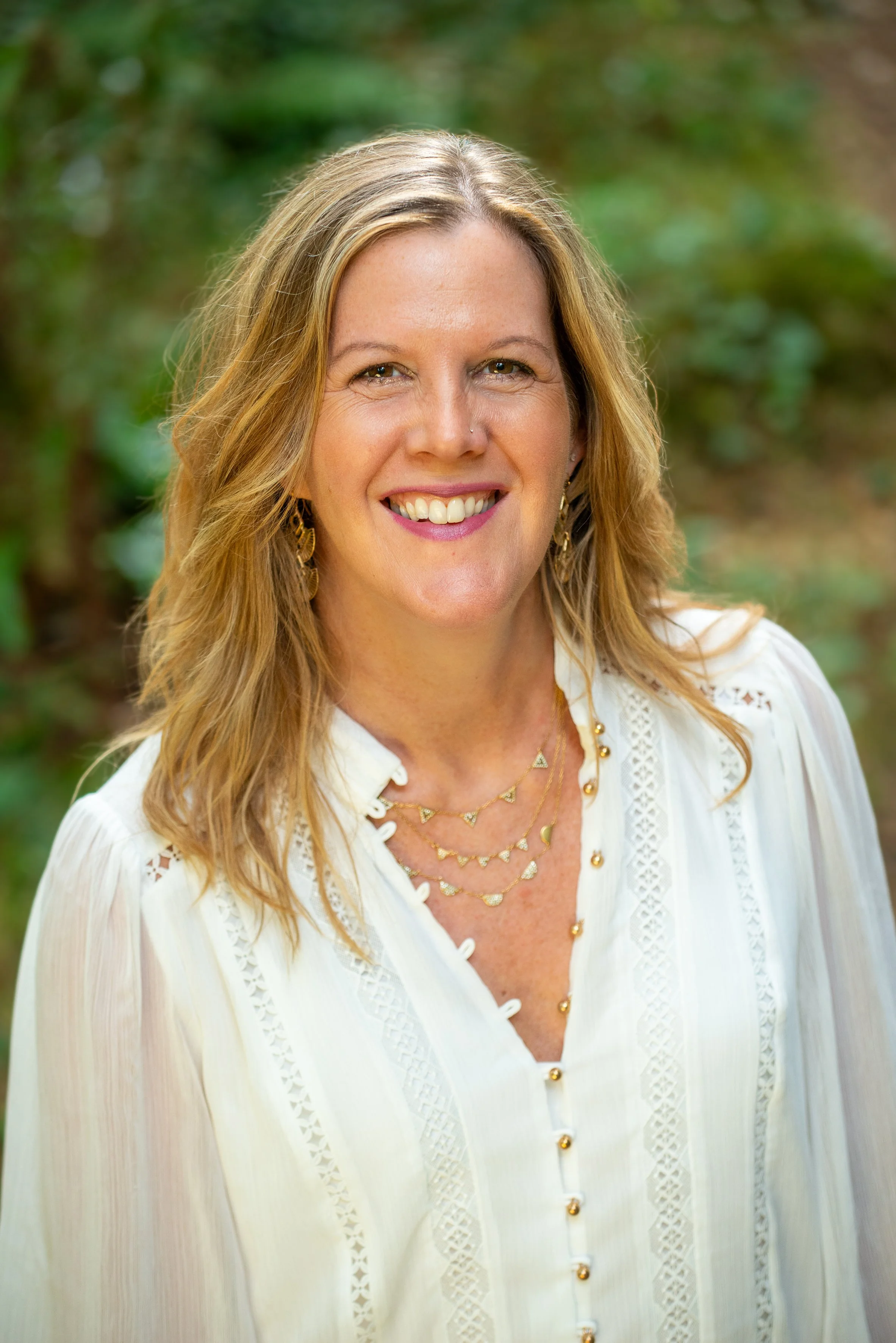 A woman with long, wavy blonde hair smiling outdoors, wearing a white blouse with lace details and layered gold jewelry.