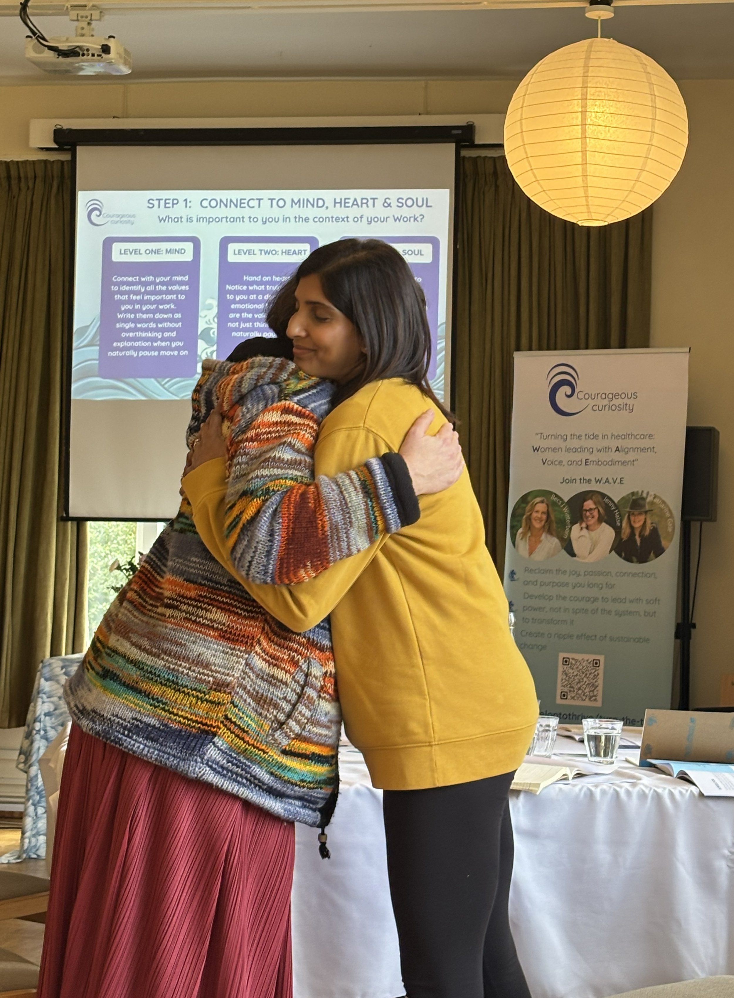 Two women hugging in a conference room with a presentation screen and a banner in the background.