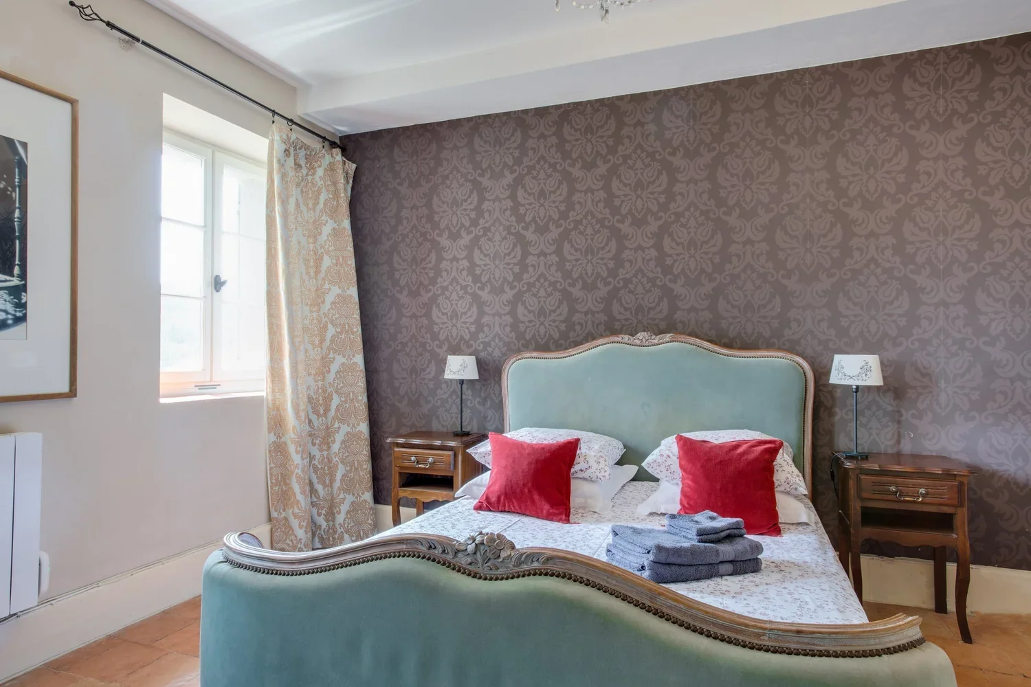 A bedroom with a vintage style, featuring a bed with a pale teal headboard and golden trim, two red pillows, a folded set of gray towels on the bed, and two wooden nightstands with white desk lamps. The wall behind the bed has brown damask wallpaper, and there is a window with white curtains and gold damask drapes on the left.