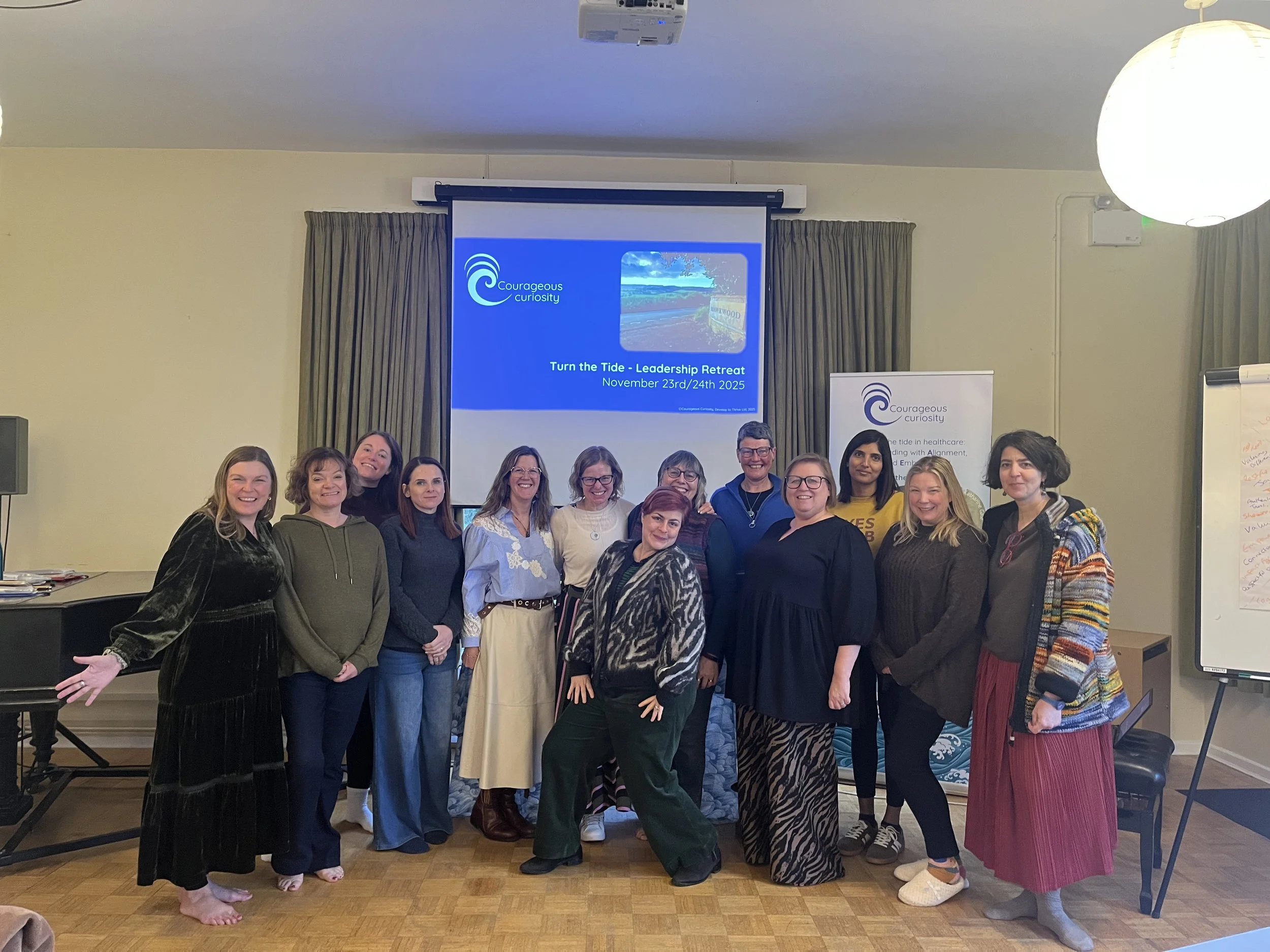 Group of women smiling and standing together in a room with a presentation screen that reads 'Turn the Tide - Leadership Retreat' and dates 'November 23rd/24th 2025'.
