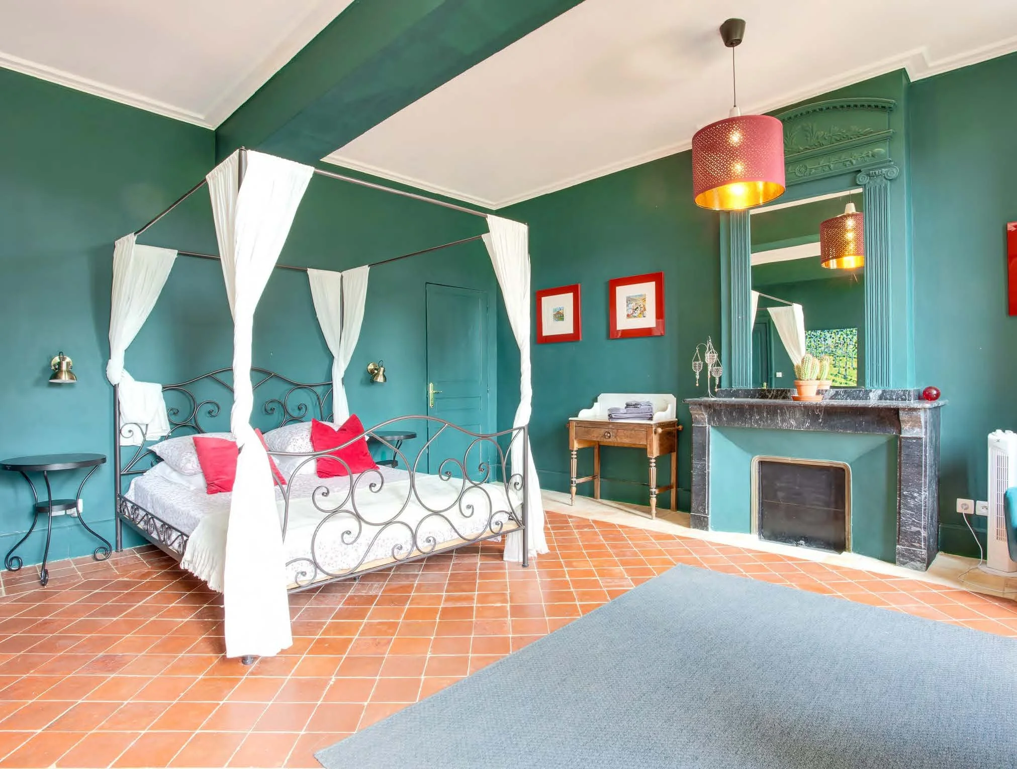 A bedroom with green walls, a metal four-poster bed with white bedding and red pillows, and terracotta tiled floors. There is a black fireplace with a black marble mantel, and a mirror with teal decorative columns. The room has pink ceiling lamps and framed artwork on the walls.