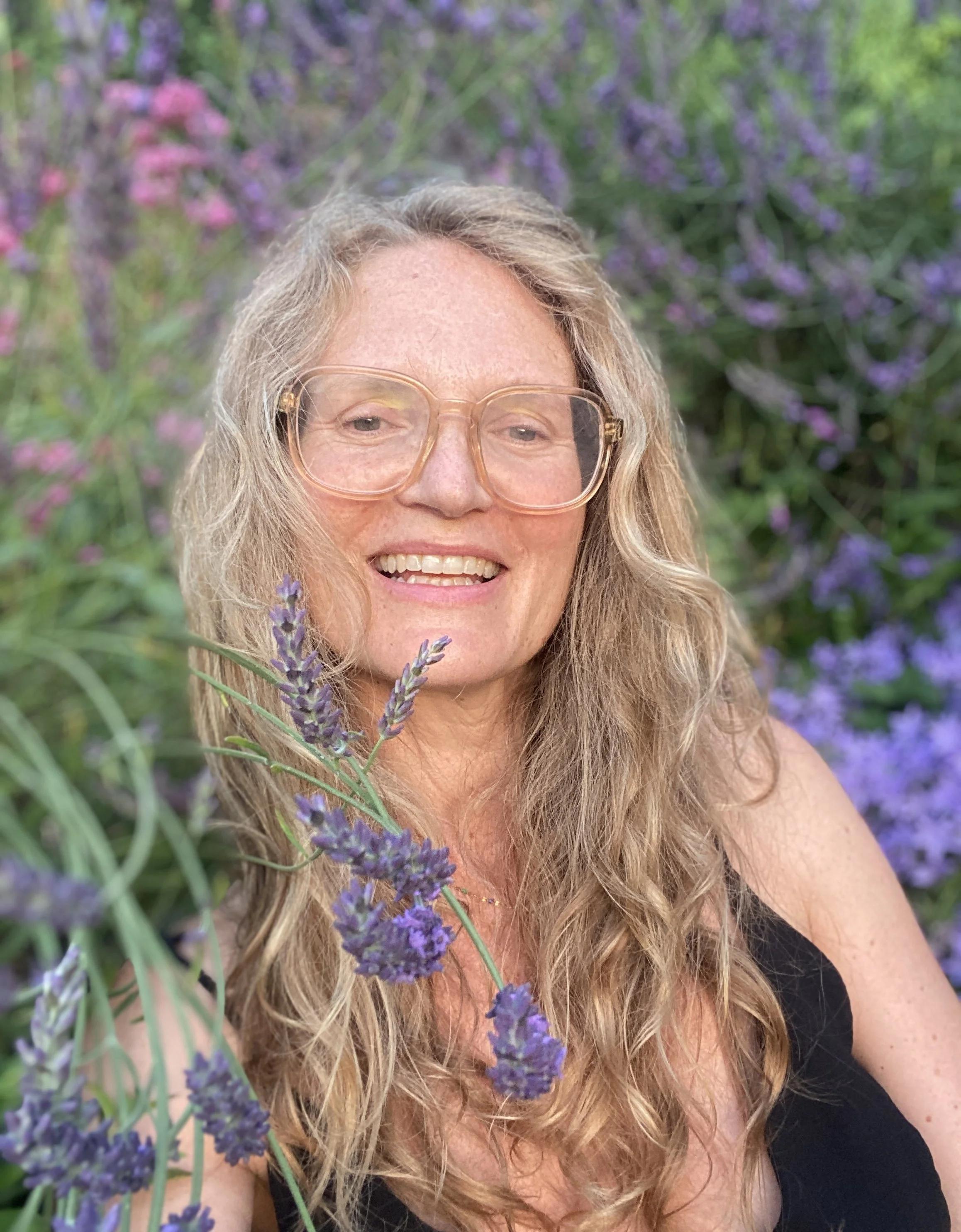 A woman with long wavy blonde hair smiling outdoors, wearing large glasses, surrounded by purple lavender flowers and greenery.