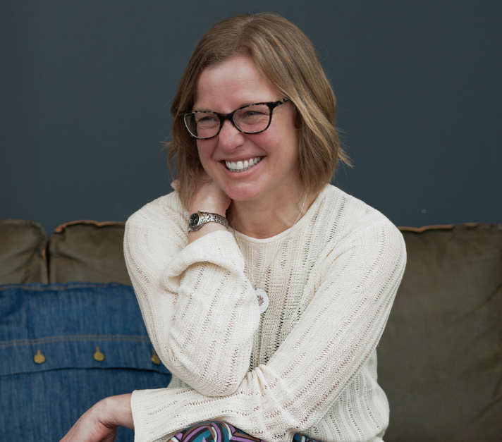 A woman with glasses and shoulder-length hair is smiling and sitting on a couch with a dark blue wall in the background.