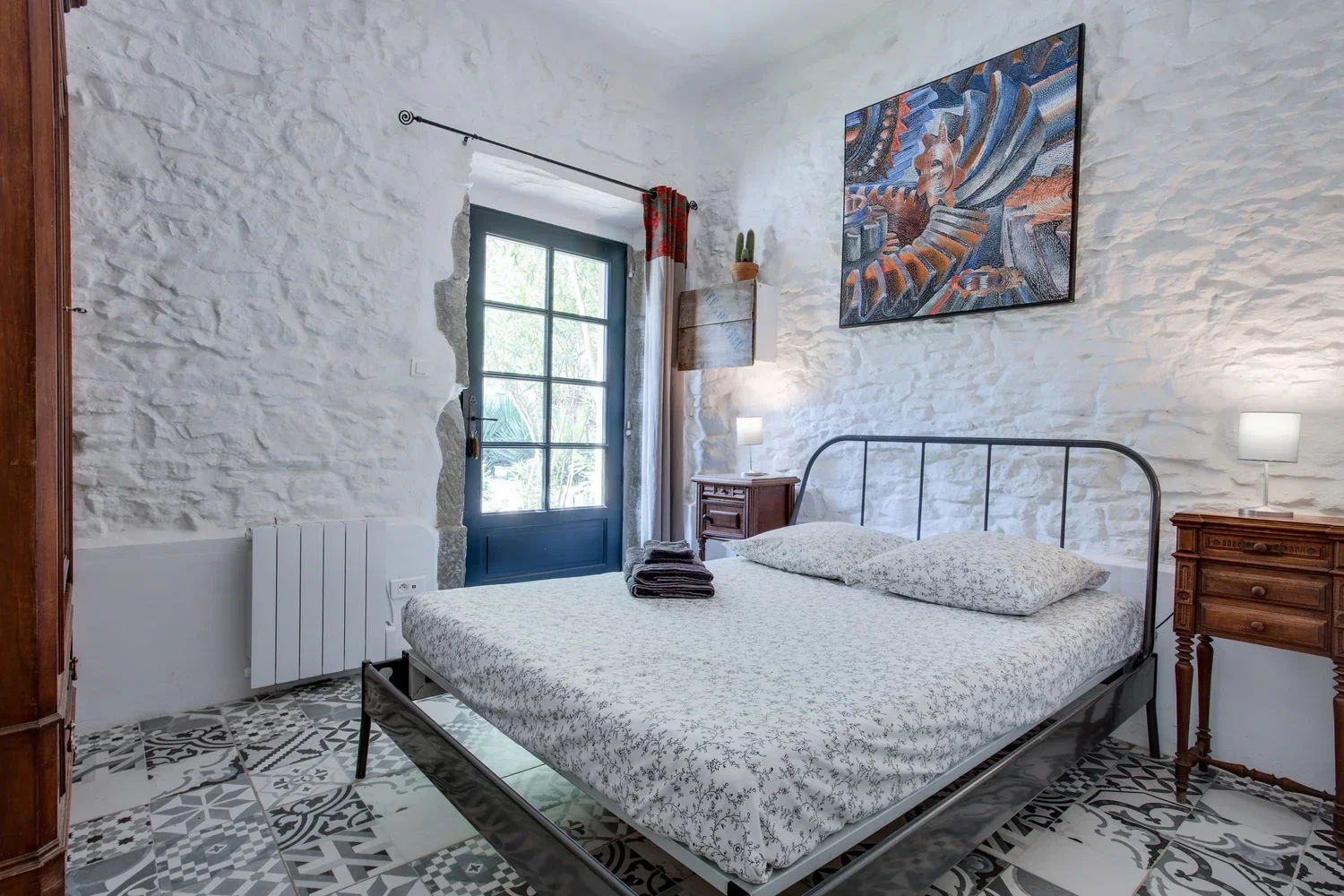 A bedroom with a black metal bed frame, white patterned bedding, two matching bedside tables with lamps, a framed colorful abstract painting, a window with curtains, and patterned floor tiles.