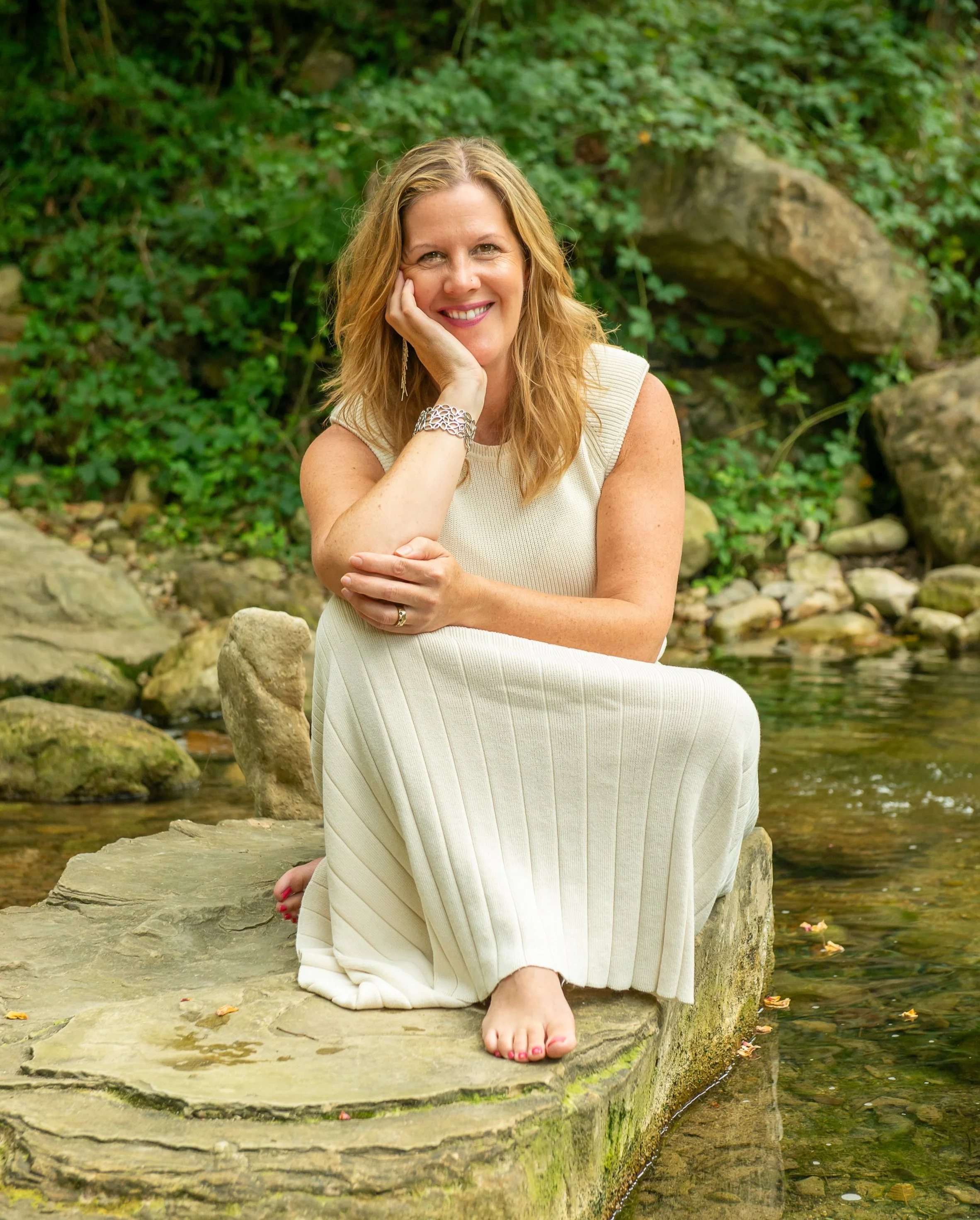A woman with long blonde hair, wearing a sleeveless cream dress, sitting on a rock by a stream in a forested area, smiling with her head resting on her hand.