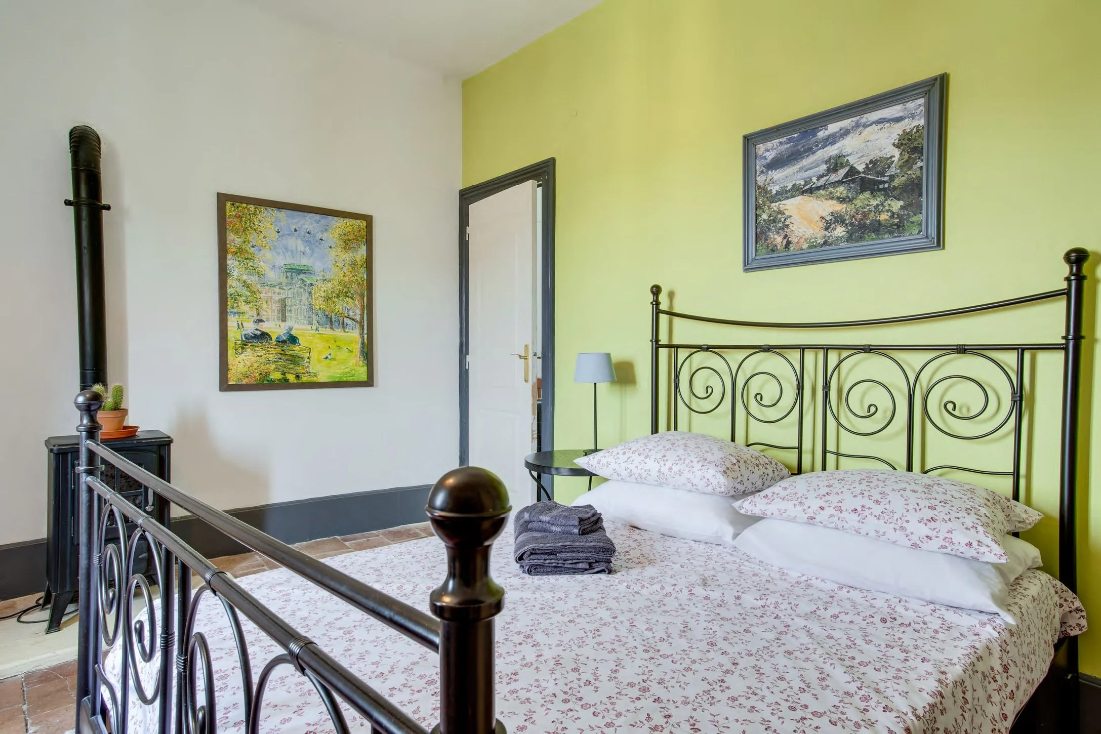 A bedroom with a black iron bed featuring floral bedding, a bedside table with a lamp, a couple of folded towels, yellow and white walls decorated with landscape paintings, and a wood stove in the corner.