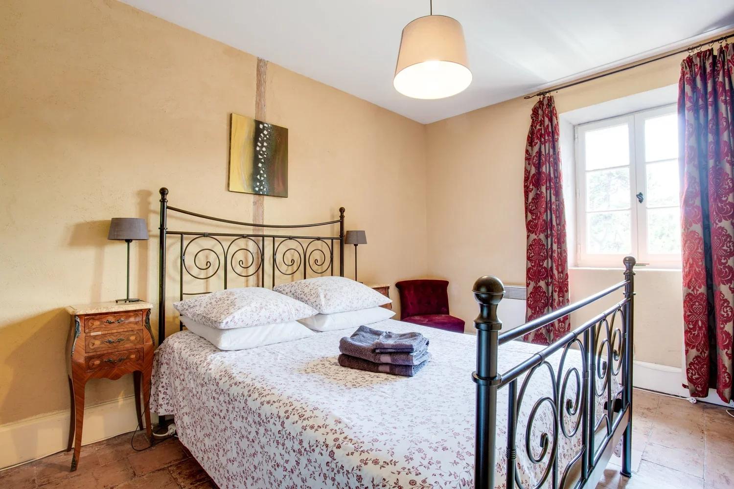 Bedroom with metal bed frame, white floral bedding, towels on the bed, wooden nightstand, table lamps, a red upholstered chair, a window with red patterned curtains, and cream-colored walls.