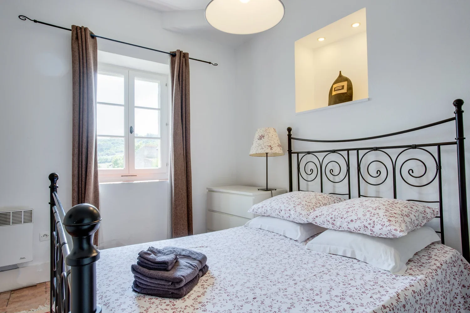 A cozy bedroom with a black metal bed frame, white bedding with a floral pattern, a window with brown curtains, a white bedside table, a floral lampshade, and a built-in wall shelf with decorative items.