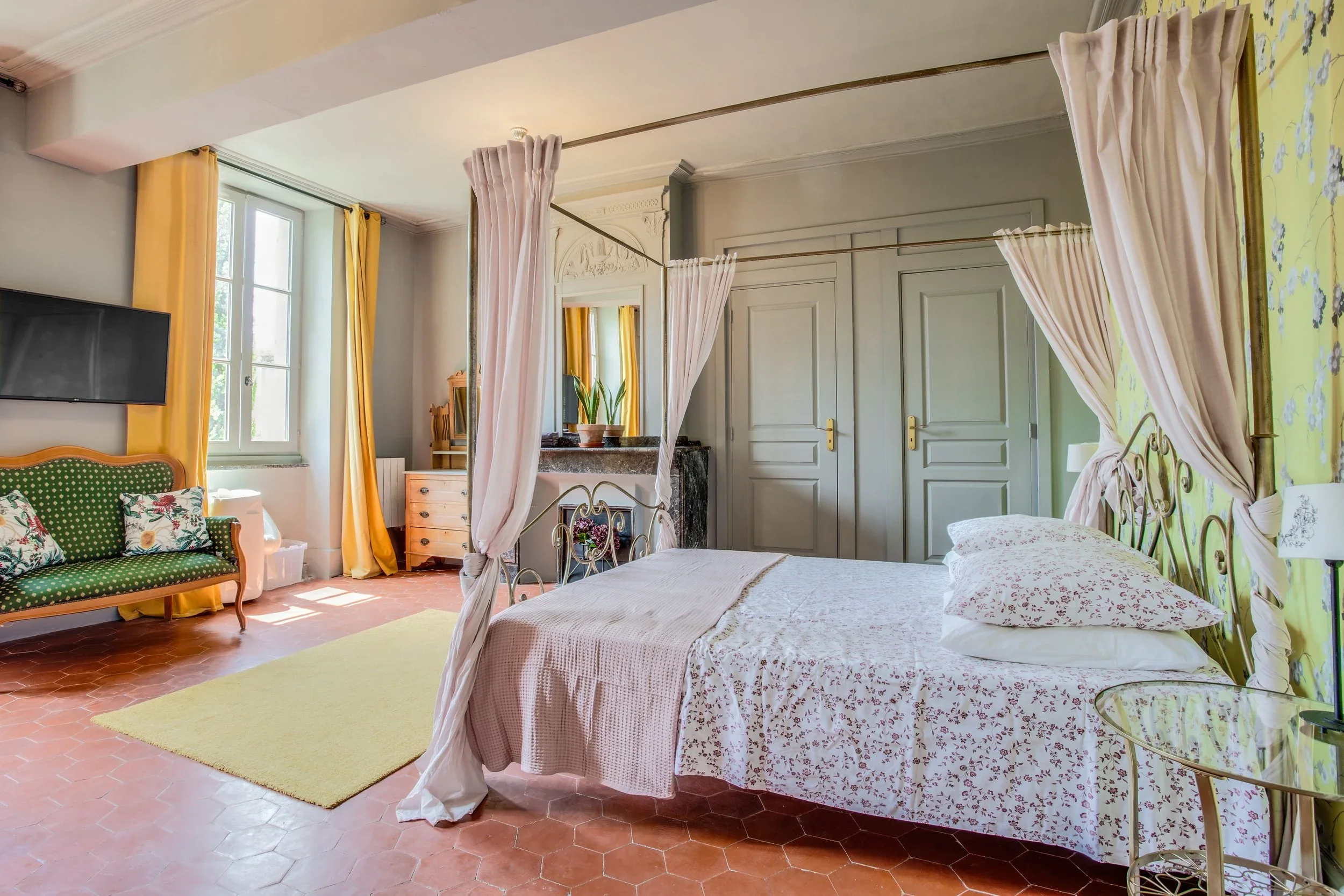 A bedroom with a four-poster bed with cream curtains, floral bedding, and pillows. There is a green velvet sofa with patterned pillows, a yellow curtain, a mirror, and a dresser near a window with yellow curtains. The room has a terracotta tile floor, a yellow rug, and a yellow floral wallpaper accent wall.