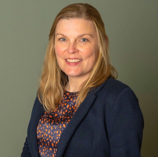 A portrait of a woman with shoulder-length blonde hair, smiling, wearing a dark blazer over a patterned blouse, standing against a plain, muted green background.