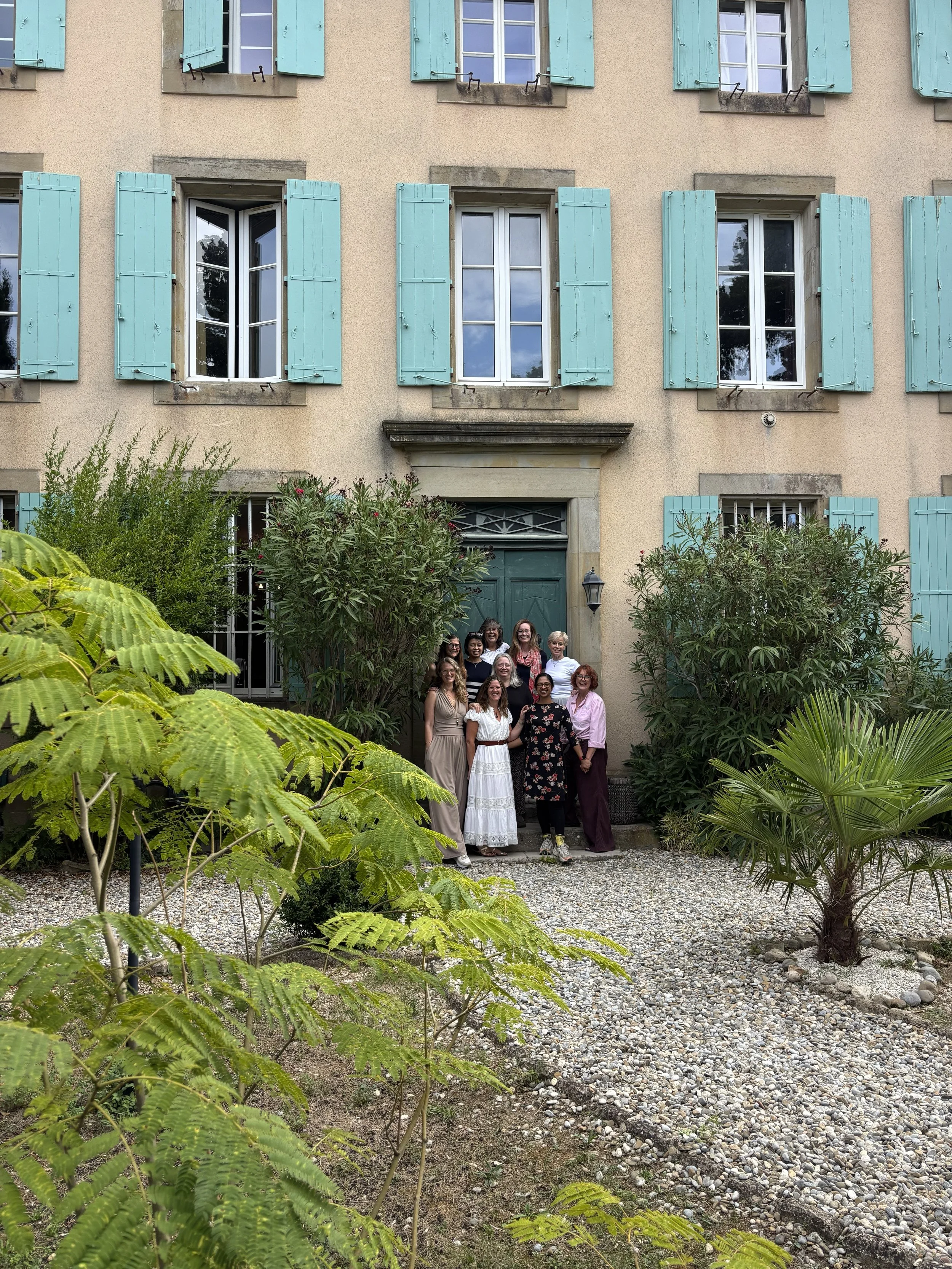 A group of ten women standing in front of a green door on a yellow building with light blue shutters, surrounded by lush bushes and a gravel pathway.