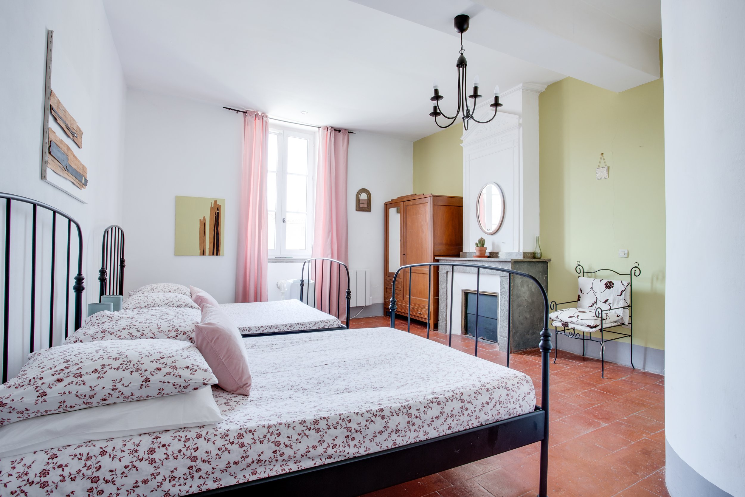 A bright bedroom with two beds, pink and white bedding, floral pillows, a wooden wardrobe, a fireplace with a small mirror, a black chandelier, a green accent wall, pink curtains, artwork on the walls, a white chair with floral cushions, a small potted cactus on the mantel, and terracotta tile flooring.