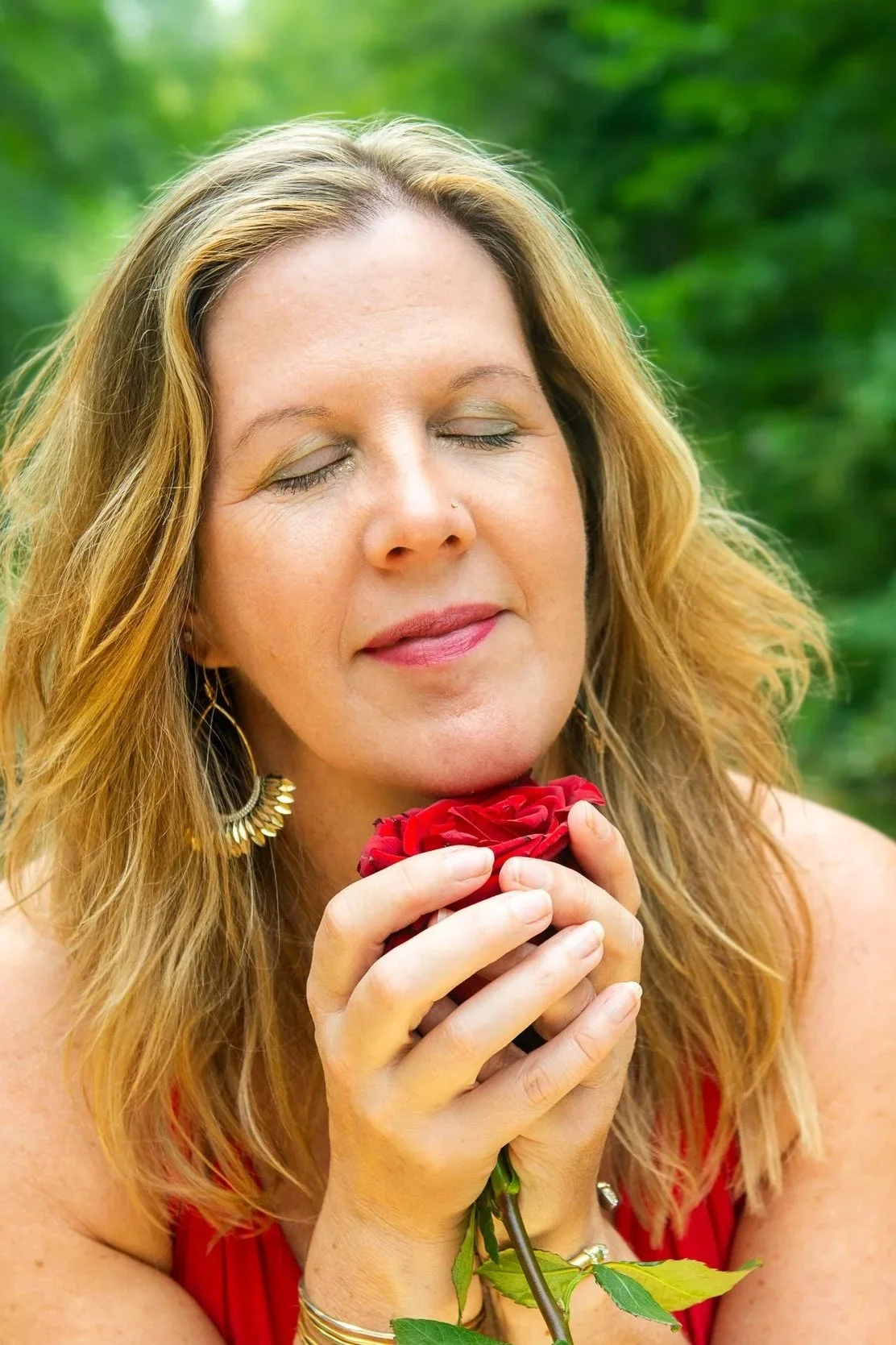 A woman with closed eyes holding a red rose close to her face outdoors.