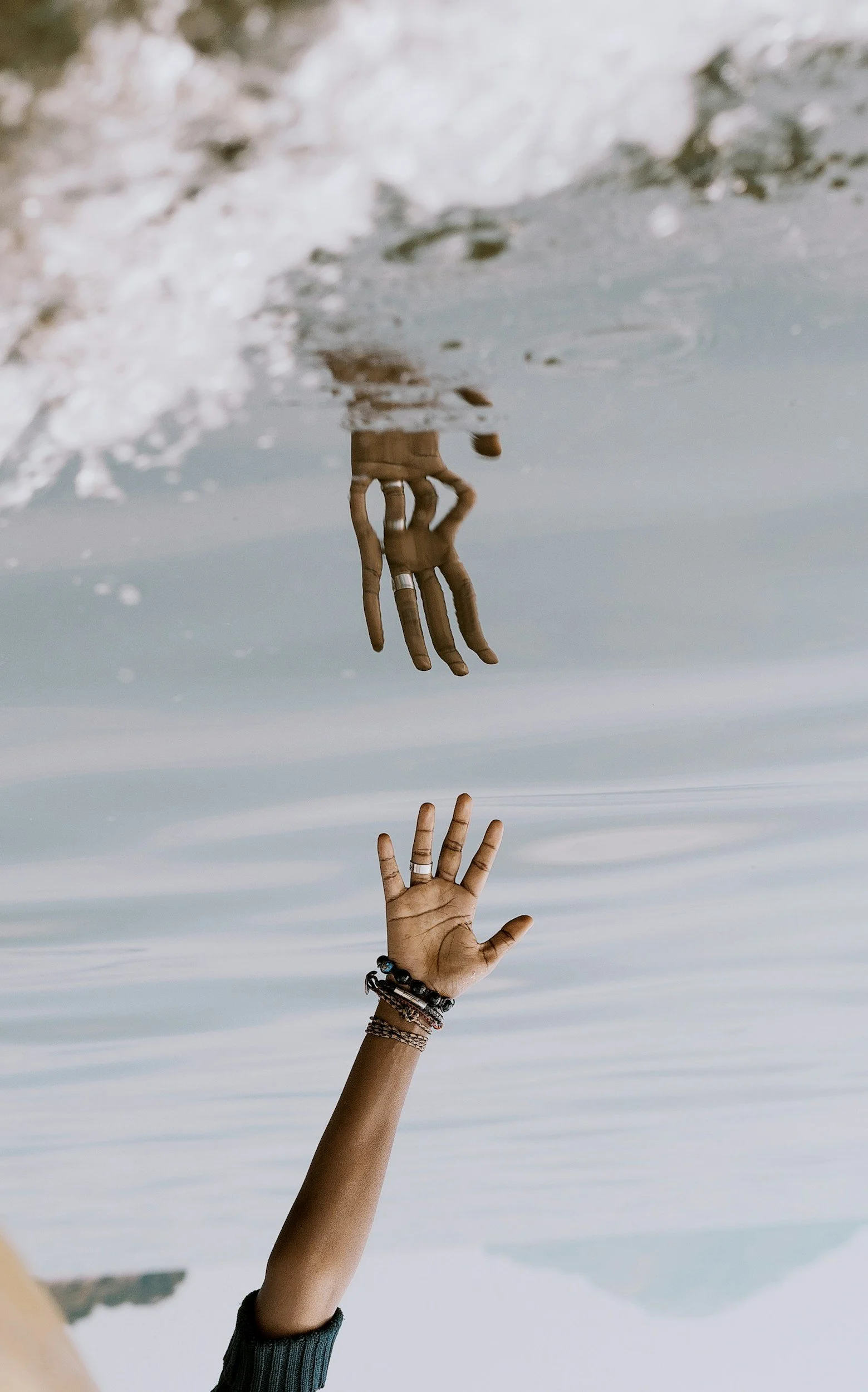 A person's hand reaching up from water near the shoreline, with mountains and sky reflected in the water.