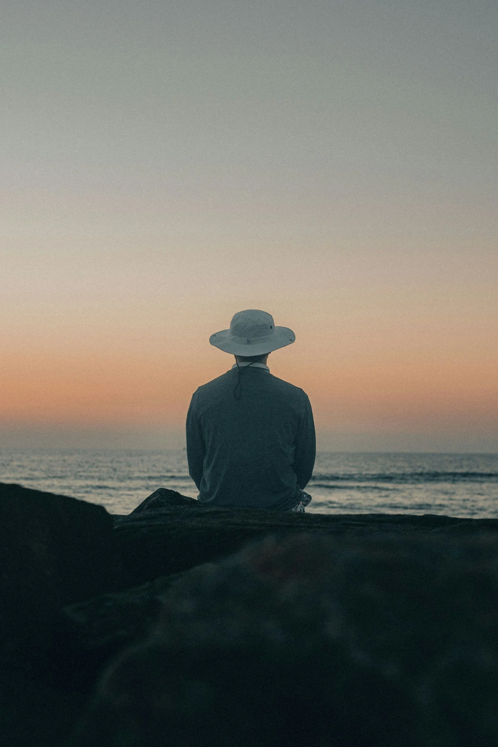 A person wearing a wide-brimmed hat and dark clothing sits on coastal rocks by the ocean, watching a calm sunset—symbolizing stillness, reflection, and inner exploration in somatic therapy