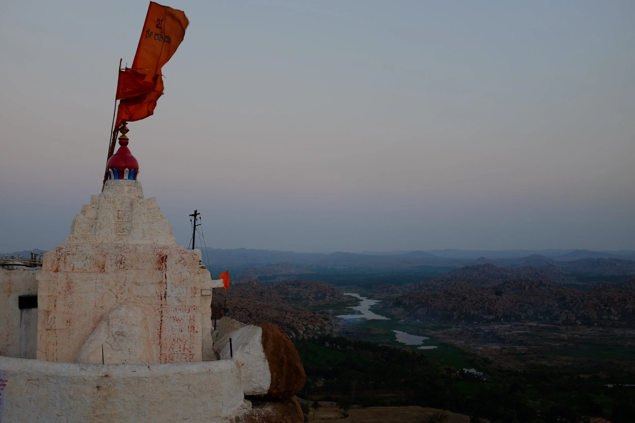 A white temple with a red, blue, and gold dome, and orange prayer flags, situated on a rocky hilltop overlooking a landscape of hills, a river, and distant mountains during dusk.