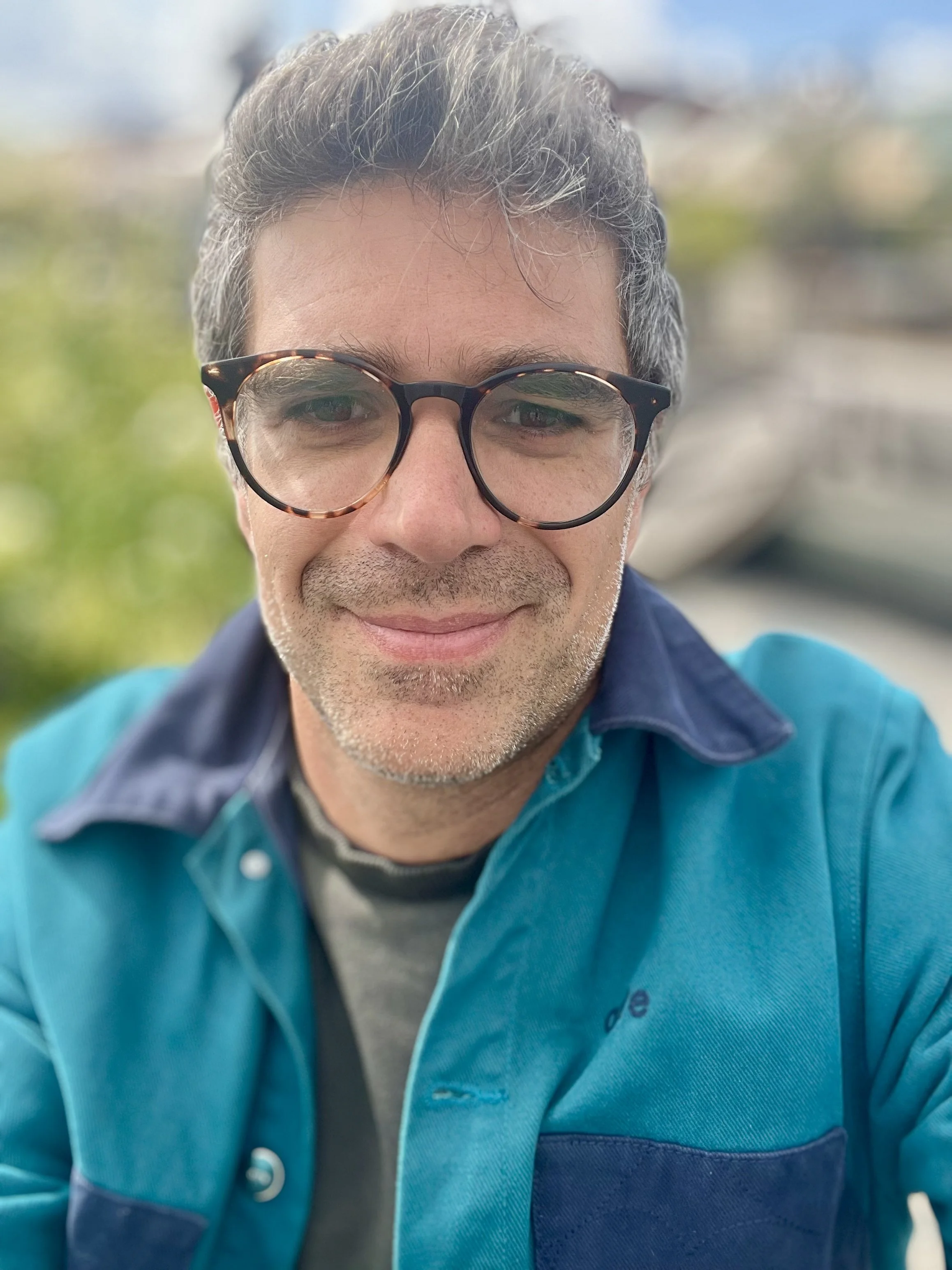 Odin, psychedelic somatic therapist, outdoors near water with trees softly blurred in the background.