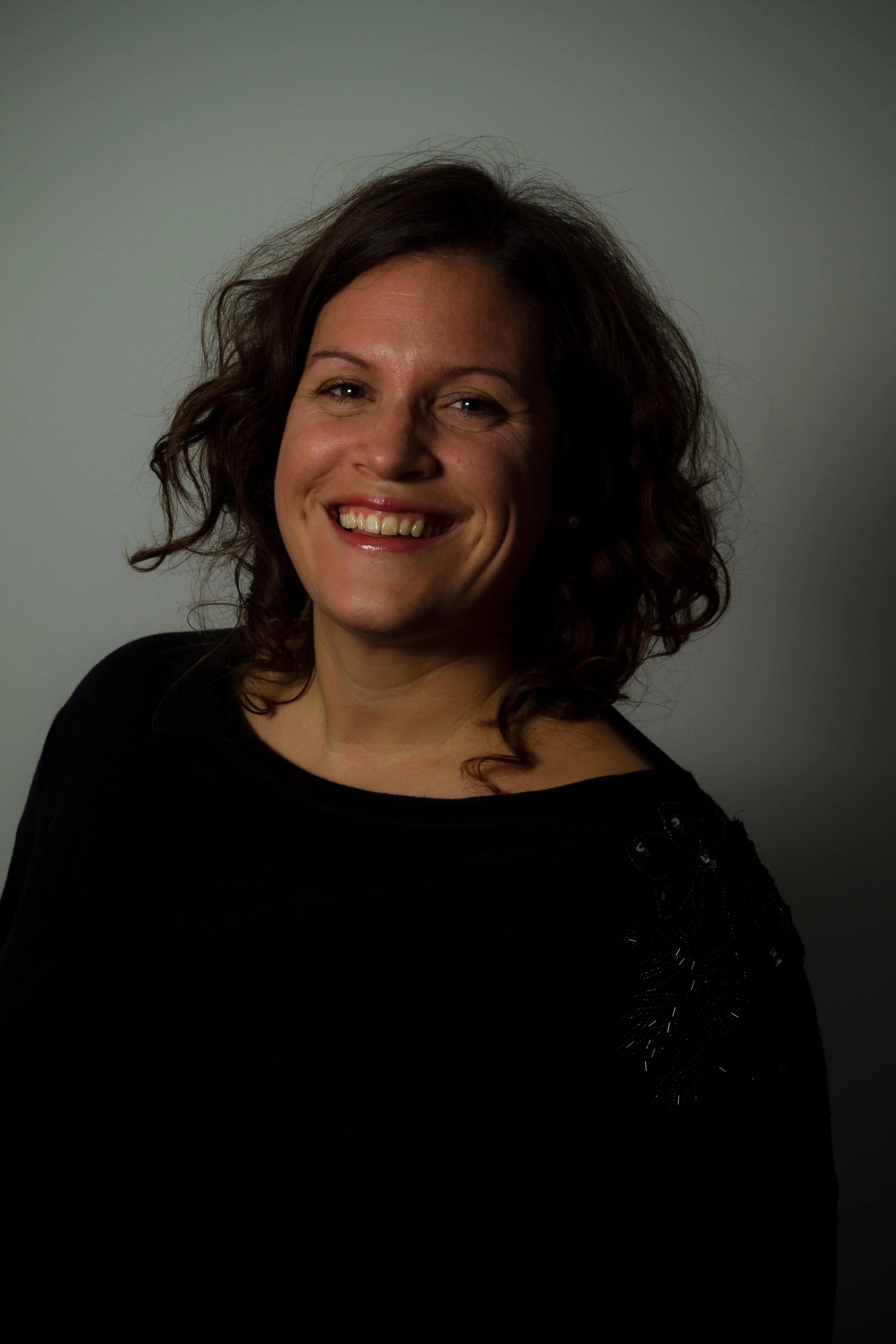 A woman with curly dark hair smiling, wearing a black top with floral embellishments on the shoulder, against a plain background.