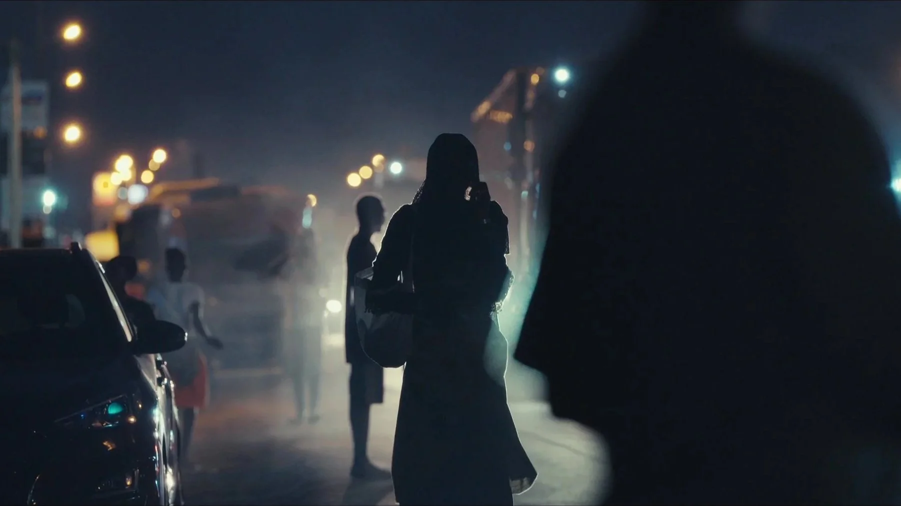 Mulher caminhando na rua à noite, carregando uma bolsa e com uma pessoa ao lado, em meio ao trânsito, com luzes de rua ao fundo.