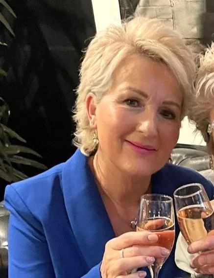 A smiling woman with short blonde hair wearing a blue outfit, holding a glass of champagne at a celebration or gathering.