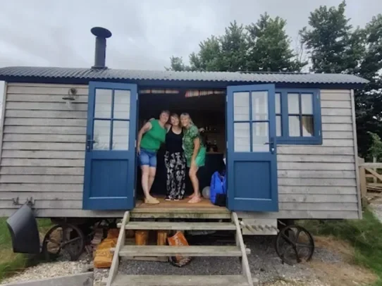 Three women standing together at the entrance of a small wooden house on wheels, with open blue doors, on a gravel and grass yard, surrounded by trees and a cloudy sky.