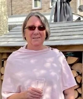 An older woman with gray hair wearing sunglasses and a pink top, holding a glass in her hand, standing outdoors in front of steps and a building.