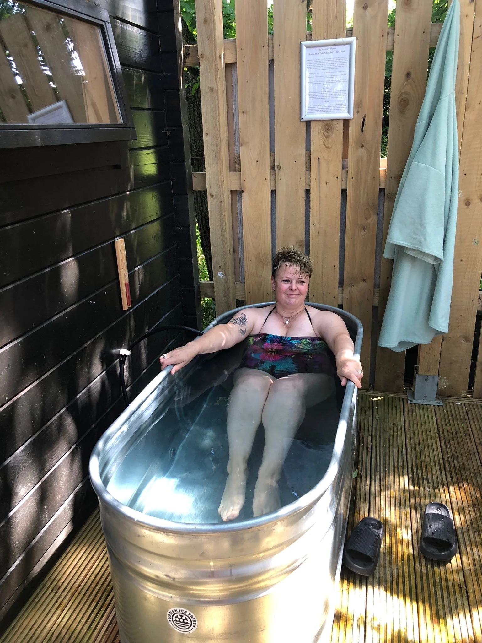 A woman relaxing in a metal hot tub on an outdoor wooden deck, with wooden fencing and a black wooden wall nearby, and a framed notice on the fence.