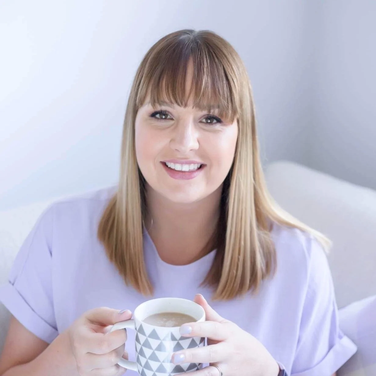 A woman with long reddish-brown hair and bangs is smiling while holding a cup of coffee or tea. She is sitting on a light-colored sofa in a bright, minimalistic room.