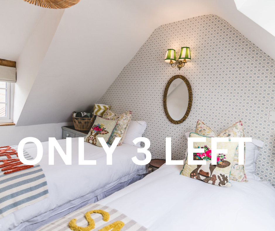 Two twin beds in a cozy attic bedroom with patterned wallpaper, decorative pillows, a mirror, a wall sconce with green lampshades, a window with a shade, and a small bedside table.