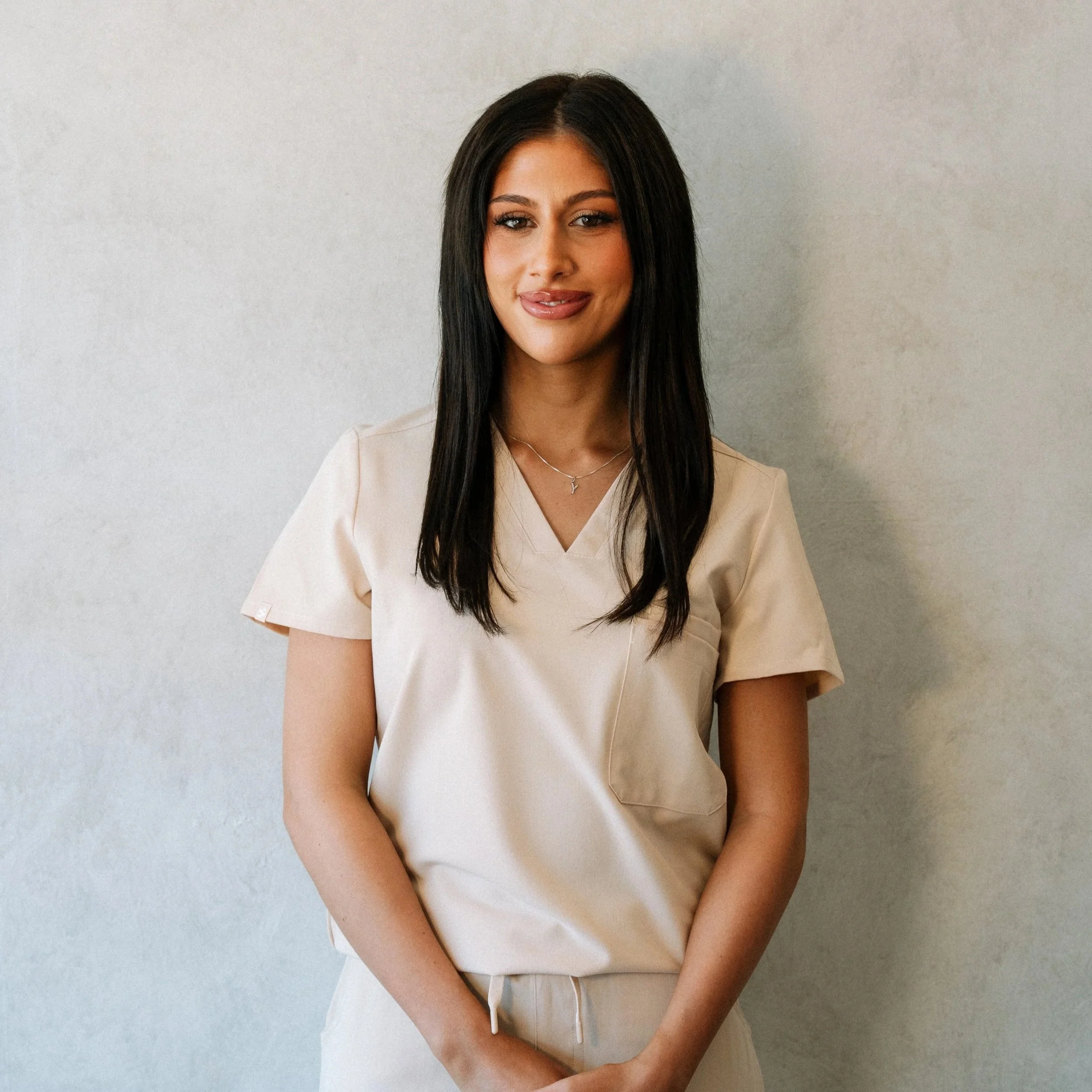 Yasmine enthoven beauty therapist. A young woman with long dark hair and light makeup, wearing a beige dress, standing against a plain light-colored wall.
