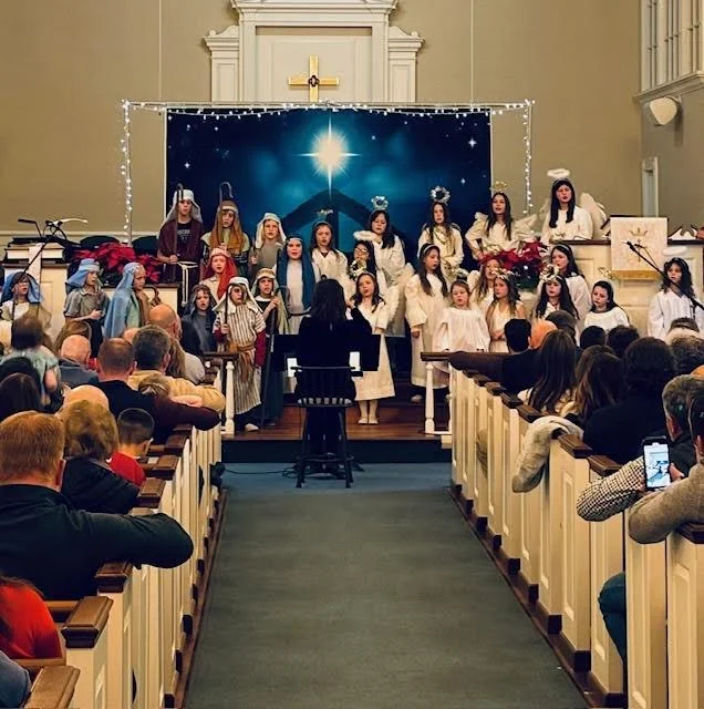 Children performing a Christmas nativity scene on stage in a church with audience watching.