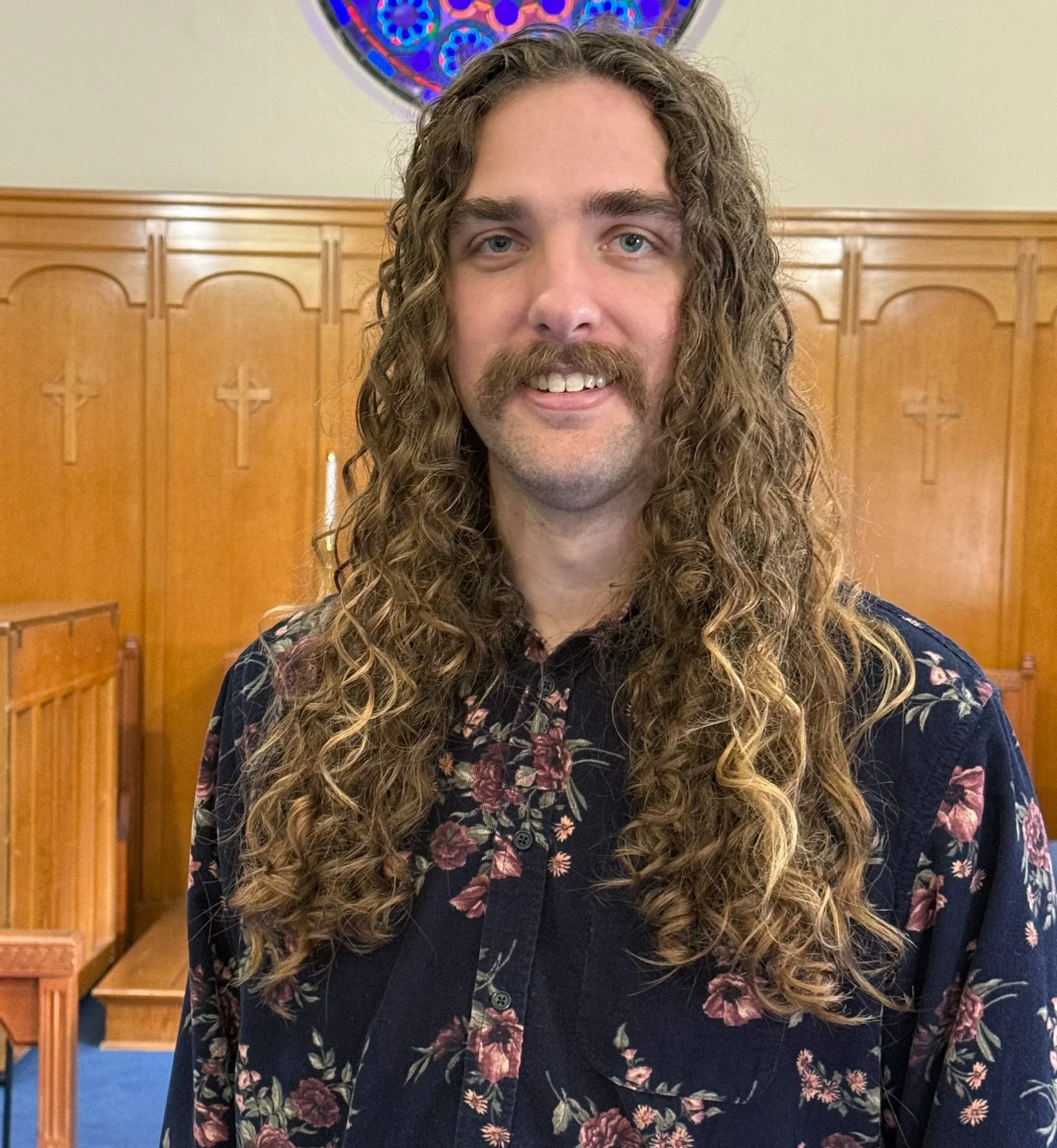 A man with long curly hair, a mustache, and a goatee smiling, standing inside a church with wooden pews and a stained-glass window in the background.