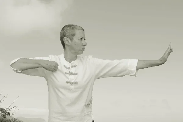 Vanda com um braço  dobrado pelo cotovelo e o outro braço esticado para o lado. O rosto virado para o lado do braço que está esticado.