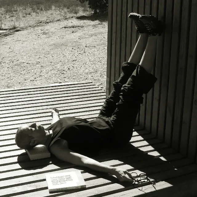 Vanda deitada no chão com um livro por baixo da cabeça e outro livro ao lado. O livro ao lado tem o título "Move your dna". As pernas estão elevadas, encostadas a uma parede de madeira. É um espaço aberto, no campo.