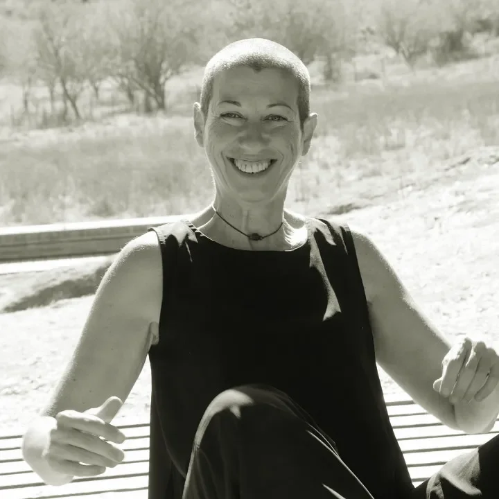 Vanda sentada no chão, num espaço em madeira. Está a sorrir, com o sol a bater no rosto. Por trás tem árvores e arbustos do campo.