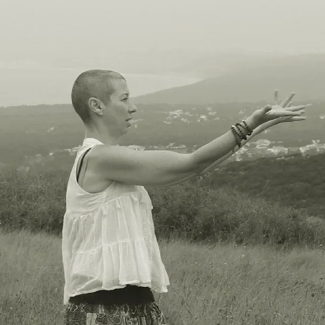 Vanda a fazer um movimento de Qi Gong, está de perfil, mostra só a parte do tronco, os braços estão estendidos à frente com as palmas para o céu, enquanto vai fechando dedo a dedo. Ao fundo vê-se o mar.