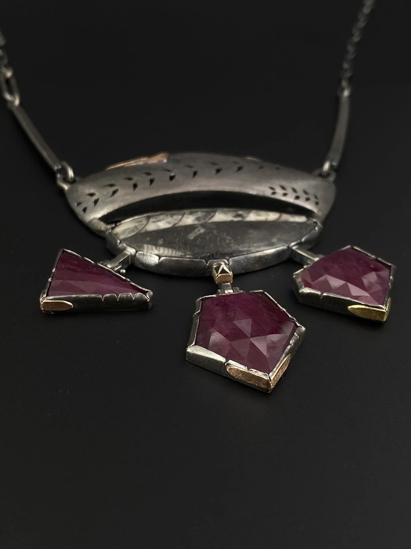 Detail view of three custom-cut red rubies in oxidized silver bezels with striking gold bottom accents, suspended from articulated hinges featuring a center black diamond.