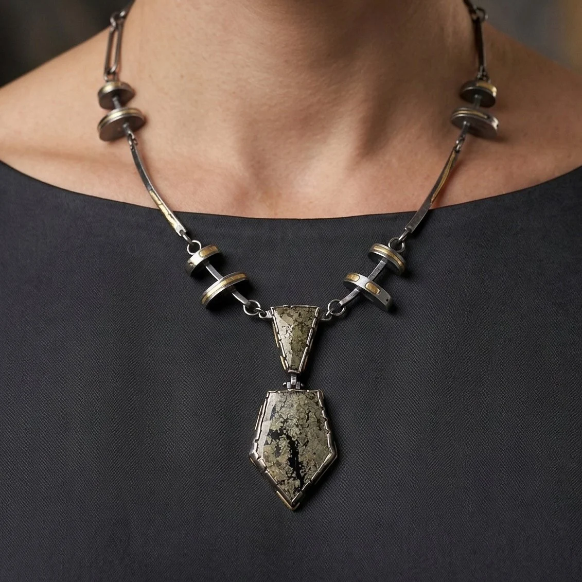Model wearing bold geometric necklace with oxidized silver links and gold accents framing a hinged chalcopyrite pendant with natural metallic gold and dark mineral patterns.